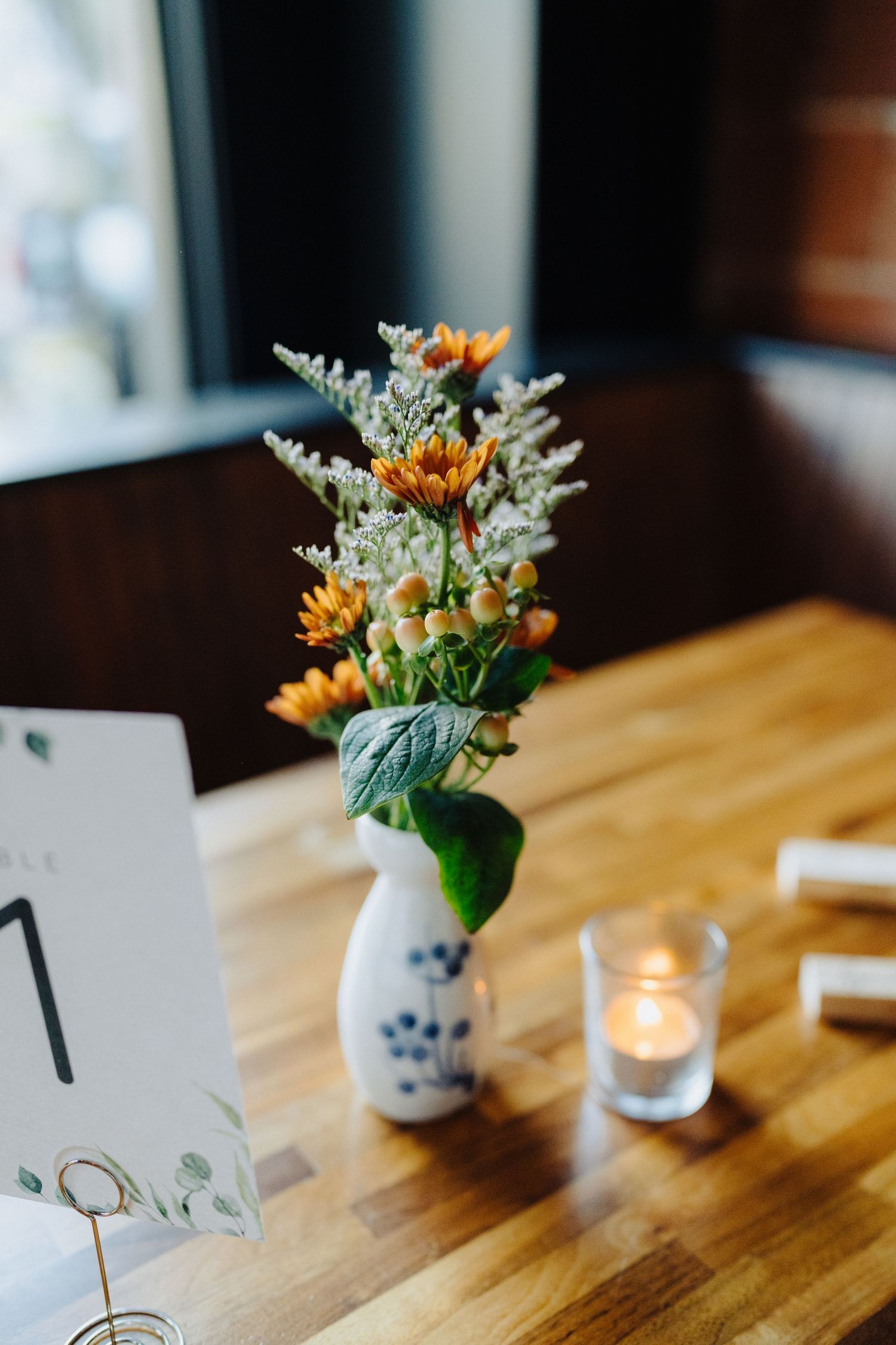 Small floral arrangement with orange blooms in a delicate blue and white vase next to table number and candle.