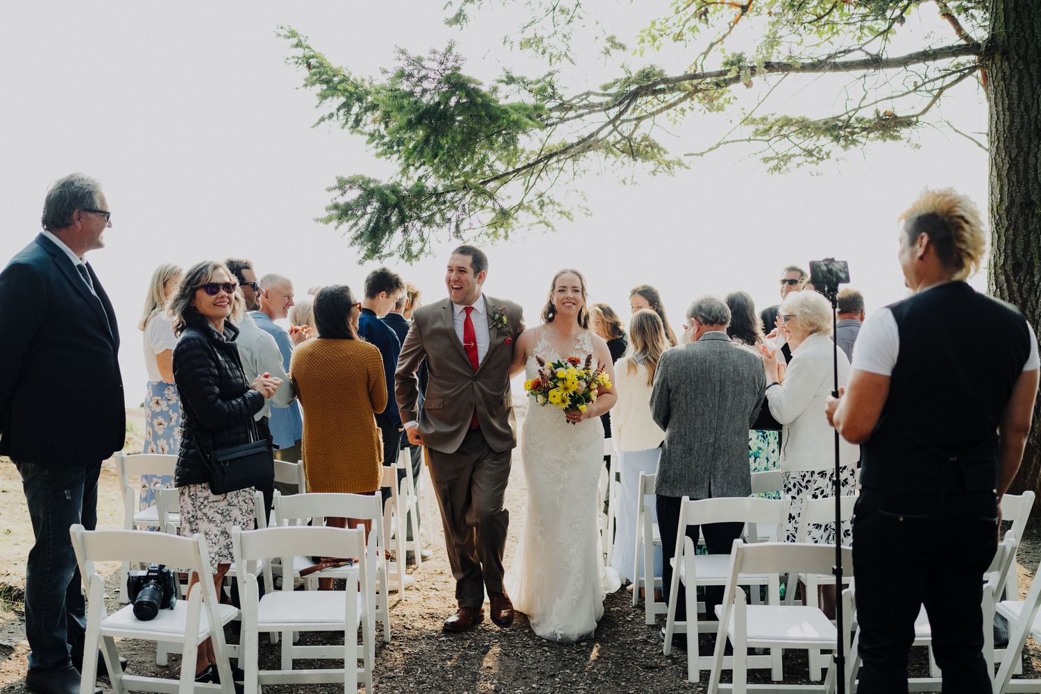 A wedding ceremony in progress with white chairs set up outdoors among evergreen trees.