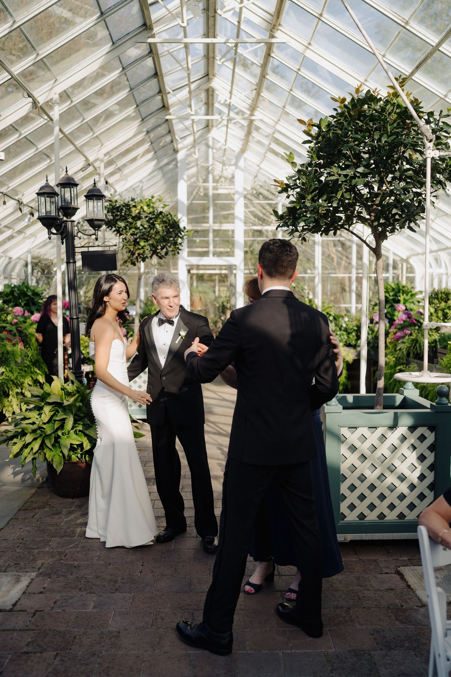 A wedding ceremony takes place in a sunlit glass conservatory decorated with potted trees and vintage lanterns.
