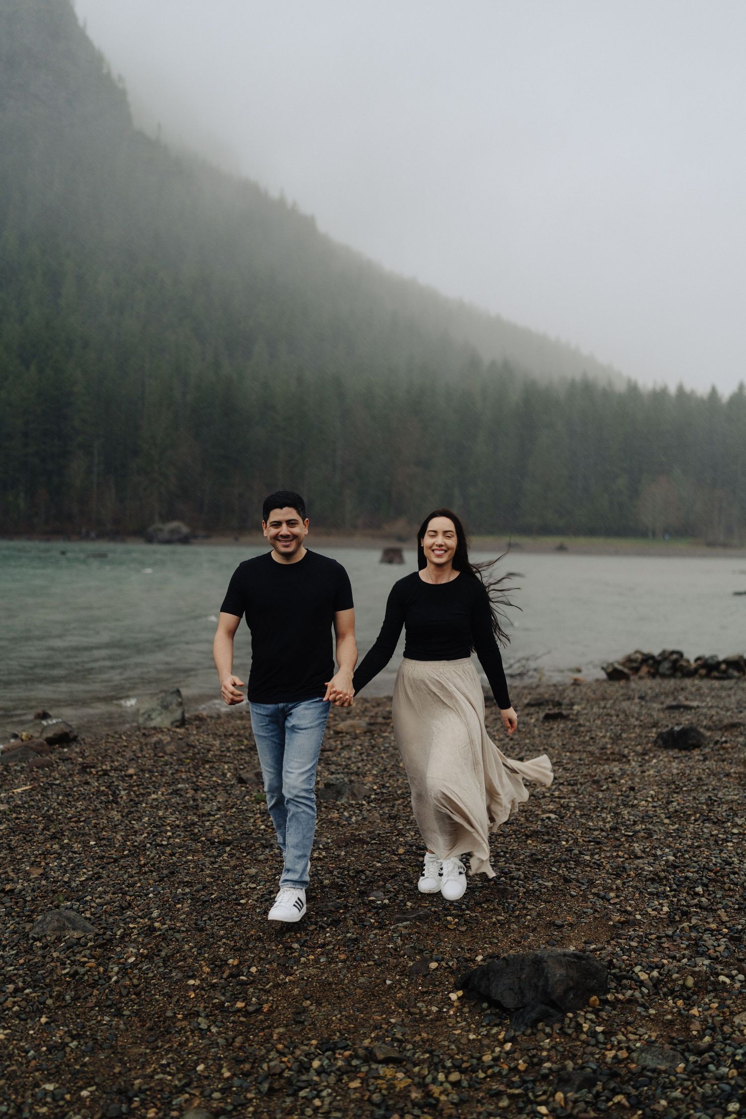 A couple strolls along a rocky beach with evergreen mountains and misty skies in the background.