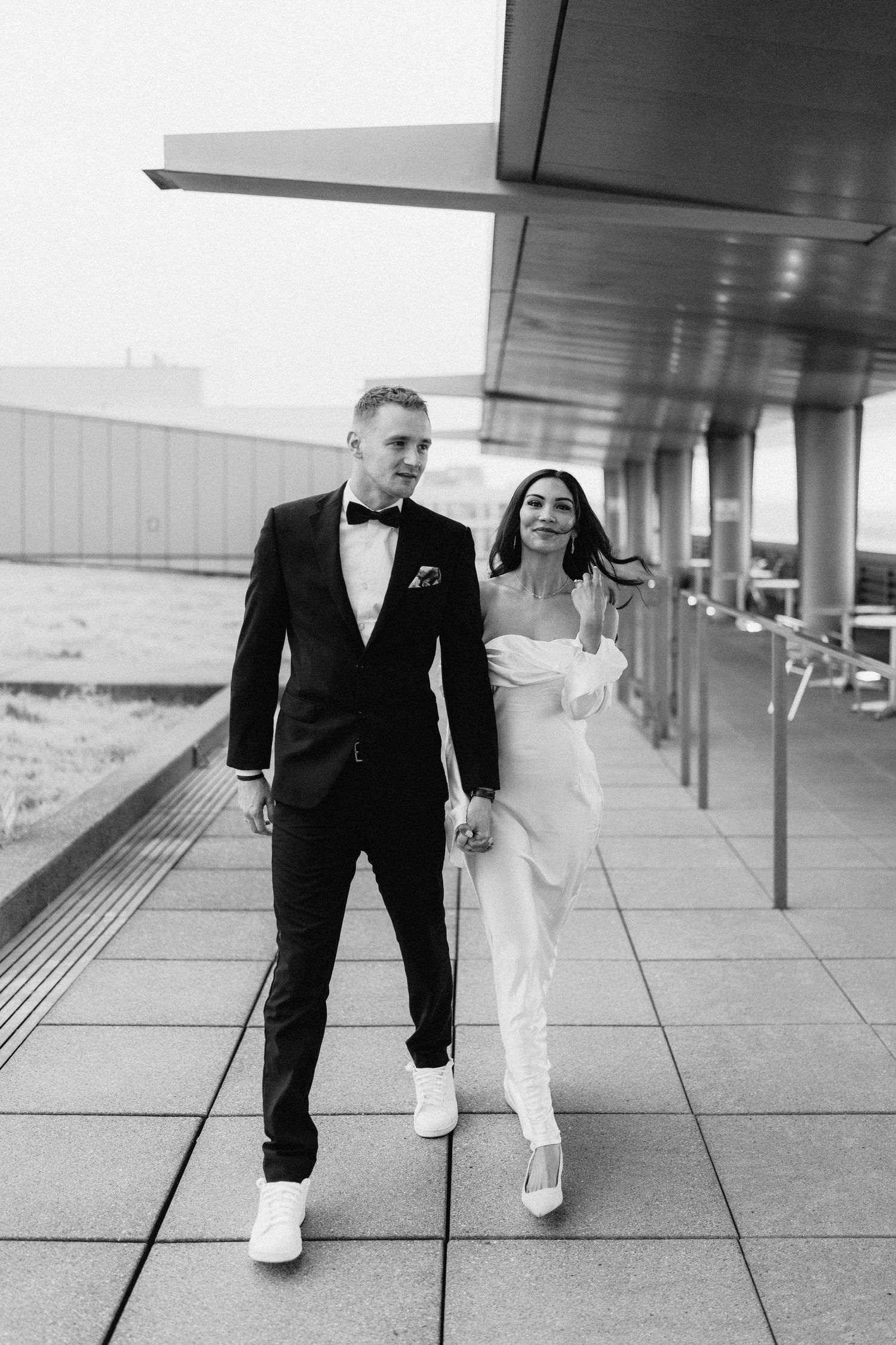 A black and white photo of a couple walking together in formal attire along a modern building exterior.