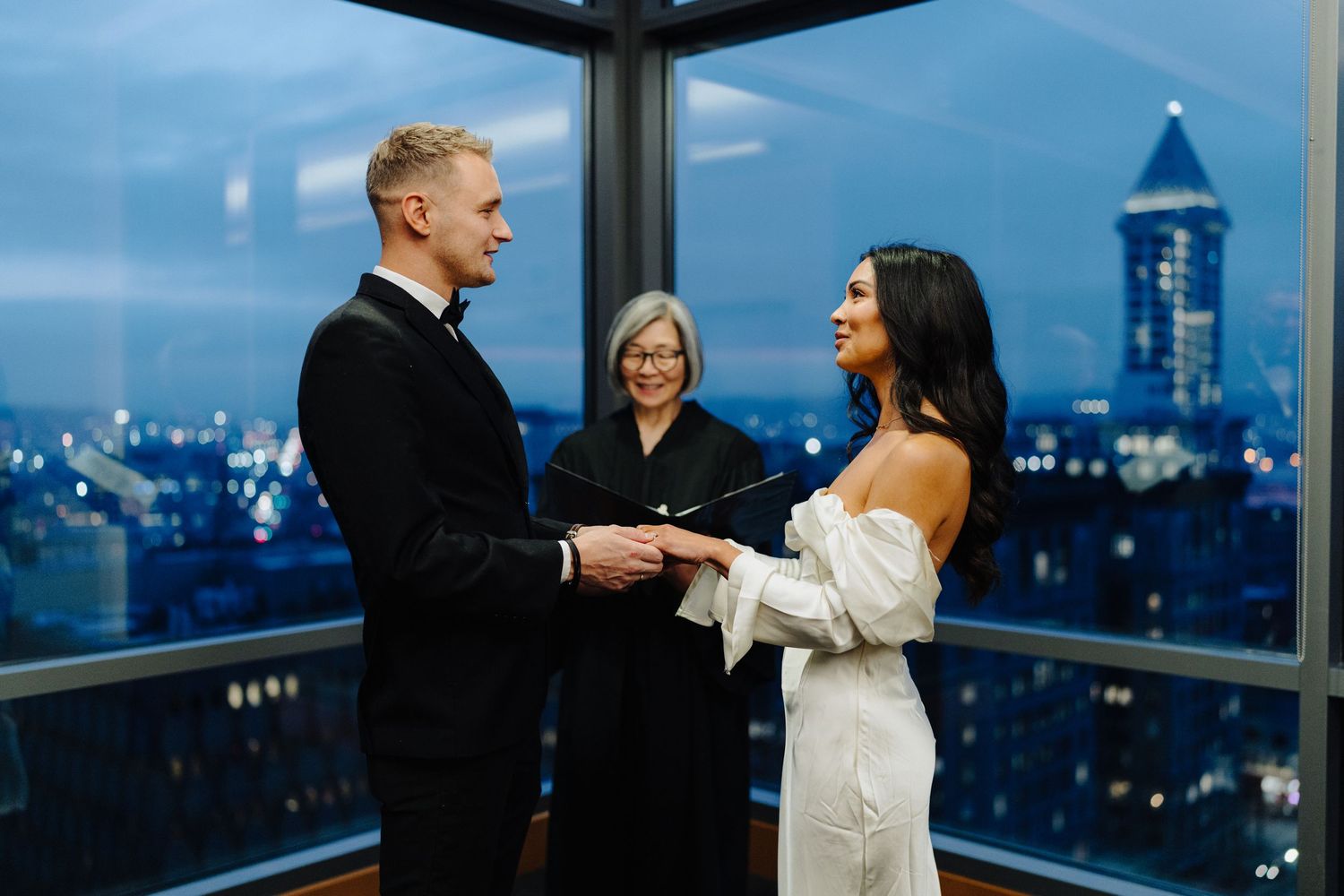 A wedding ceremony taking place in a modern high-rise with city lights visible through windows.