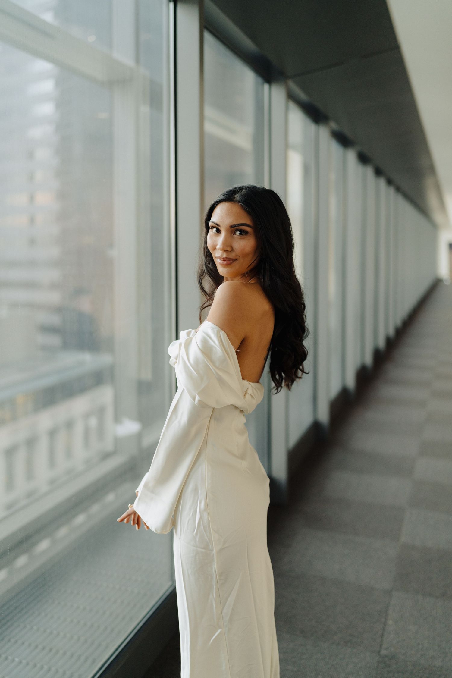 A woman in a white off-shoulder dress poses elegantly by a window in a modern hallway.