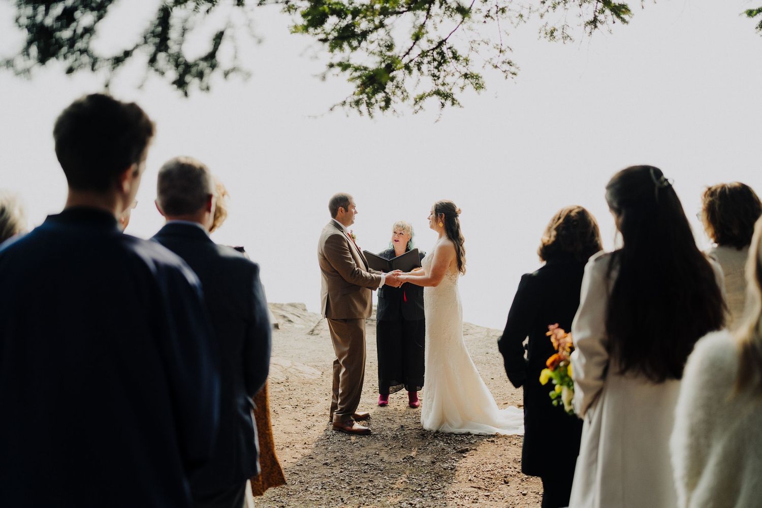 An intimate outdoor wedding ceremony takes place on a misty clifftop with guests watching the couple exchange vows.