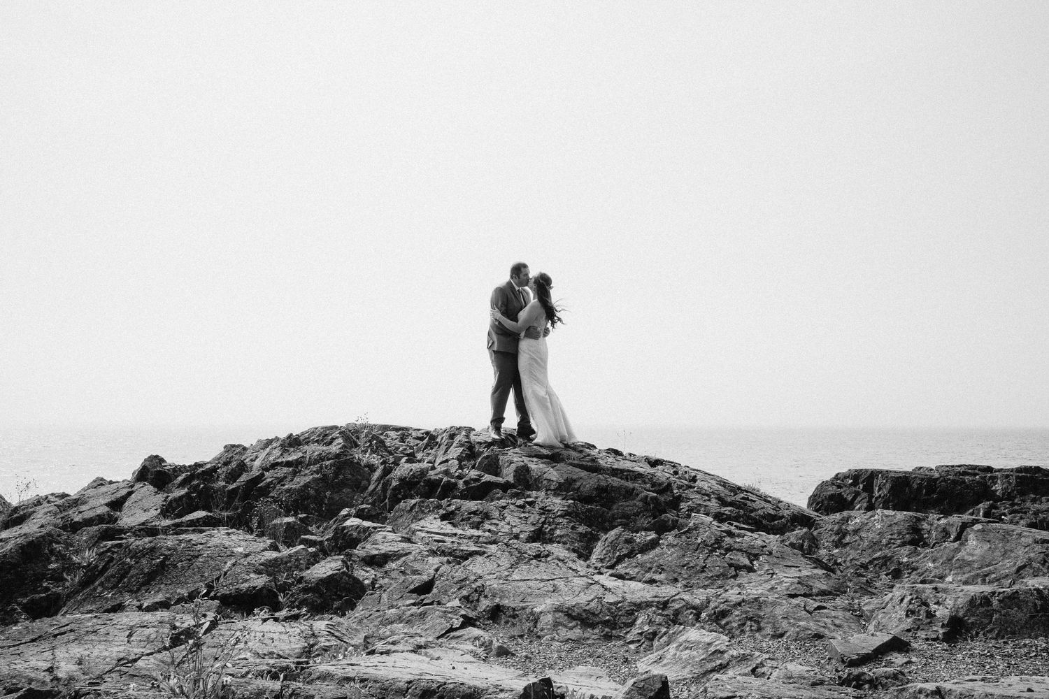 A couple embraces while standing on rocky cliffs against a misty monochrome sky.