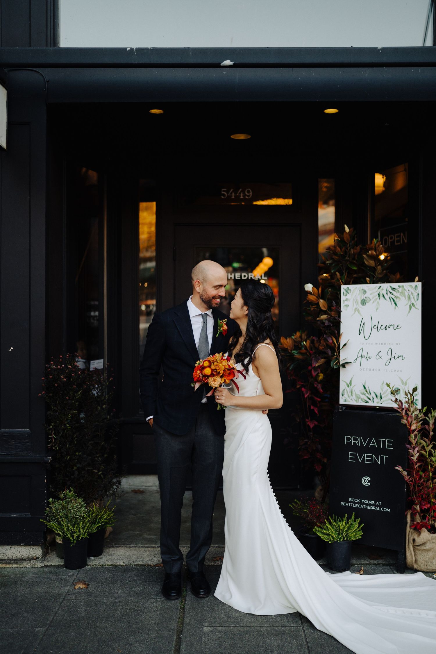 A wedding couple poses outside a venue entrance with greenery and signage.