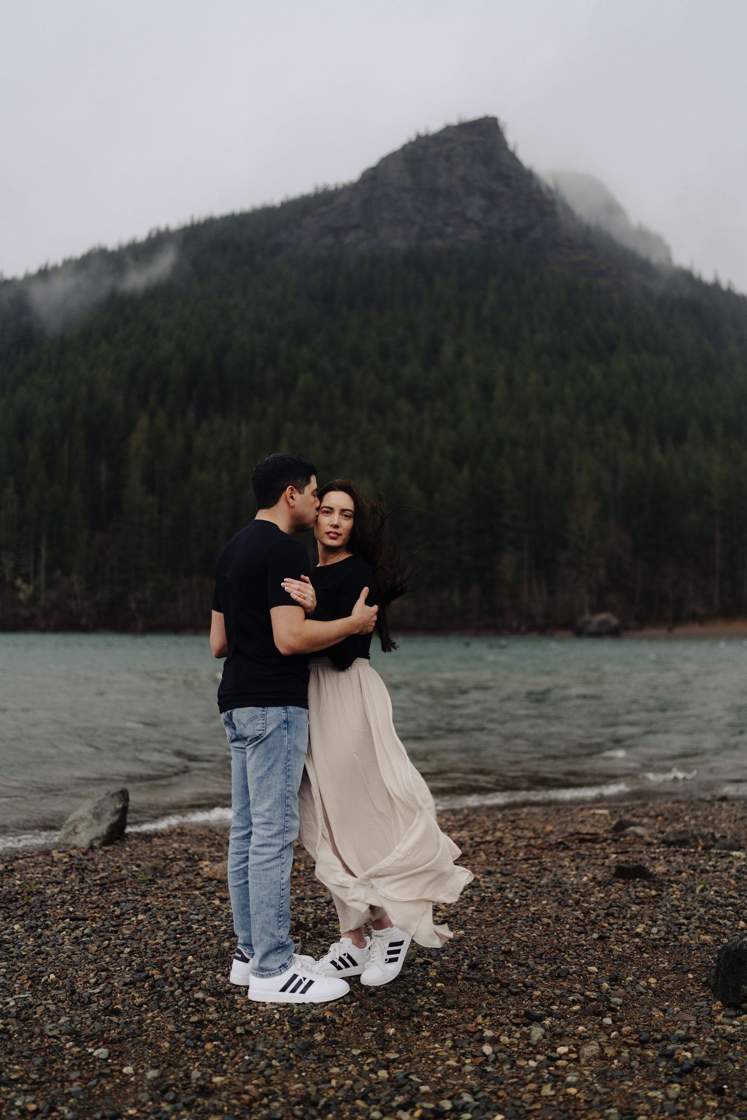 Couple sharing romantic moment on pebble beach with foggy mountain peak behind them.