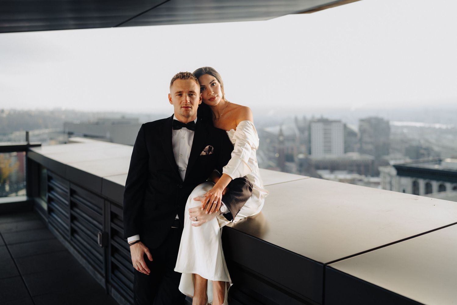 Formal couple poses against modern architectural backdrop on high-rise balcony.
