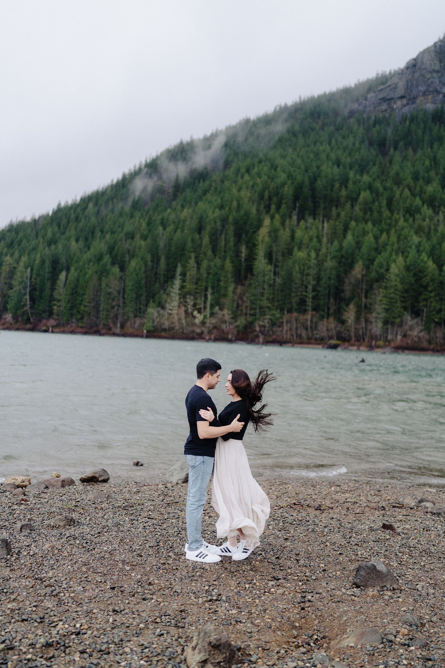 Couple dancing joyfully by lake with evergreen forest and mountain backdrop.
