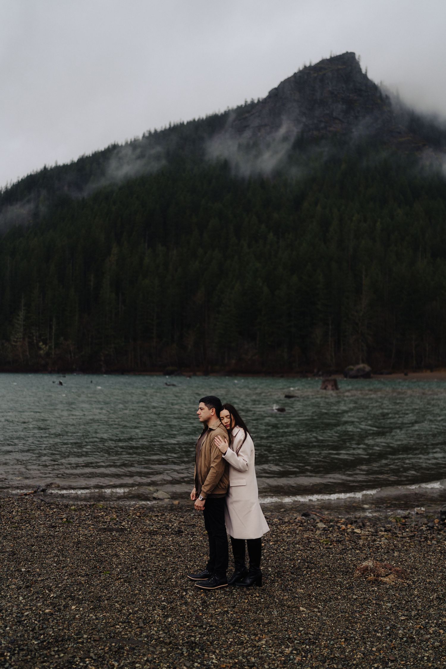 A couple stands close together on a rocky beach with misty mountain peaks looming behind.