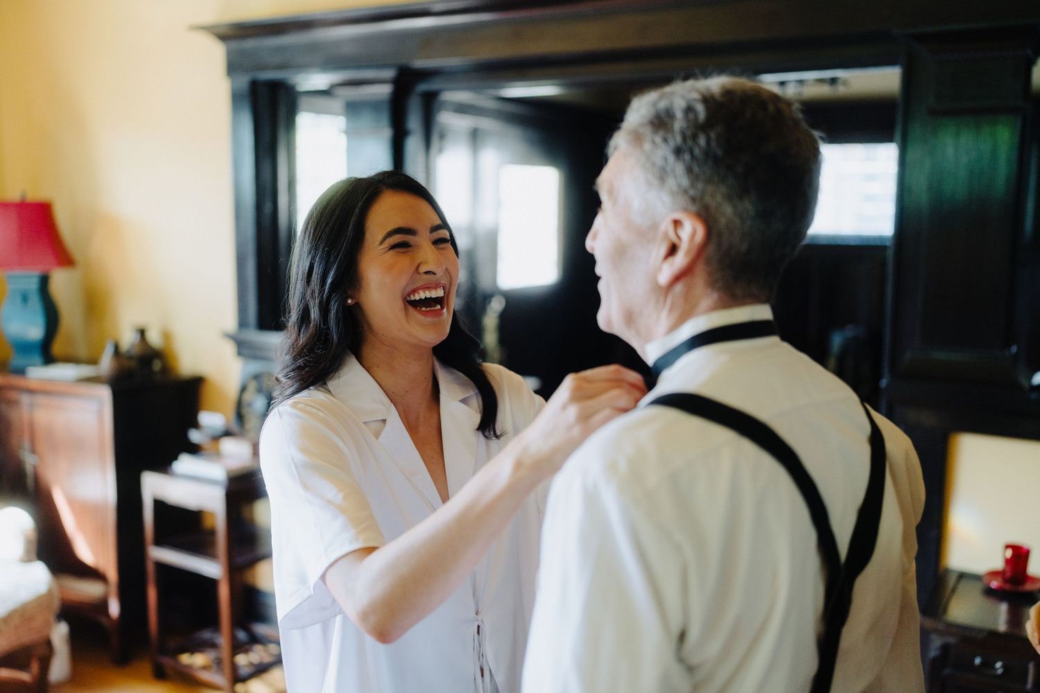 A joyful moment shared between a woman in a white robe and a man in suspenders during wedding preparations.