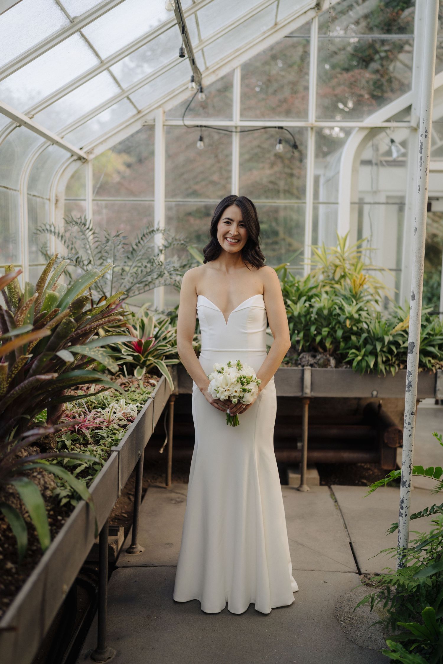 A elegant bride posing in a greenhouse filled with tropical plants and natural light.