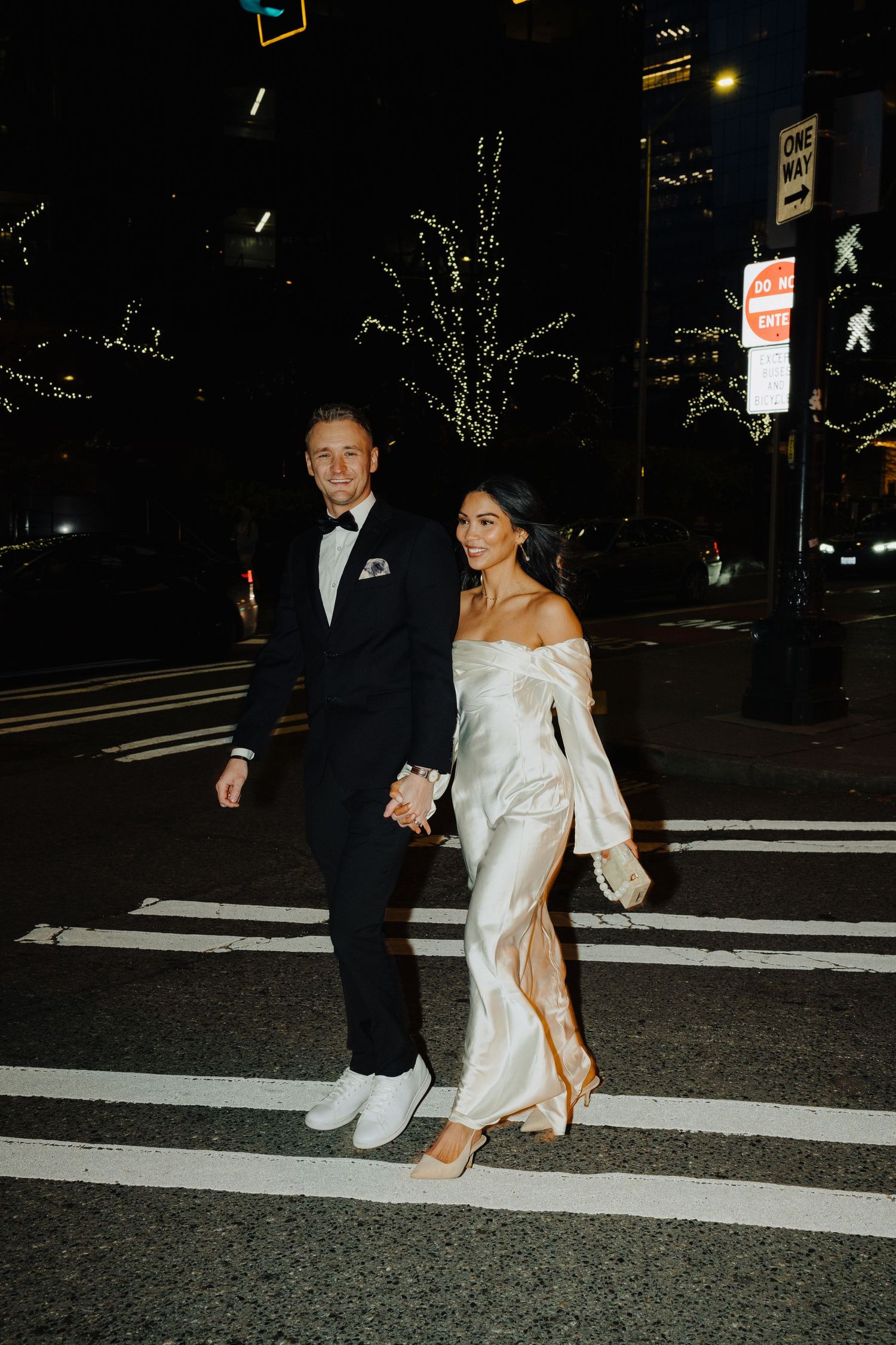 A couple in formal attire walks across an urban crosswalk with decorative lights in trees behind them.