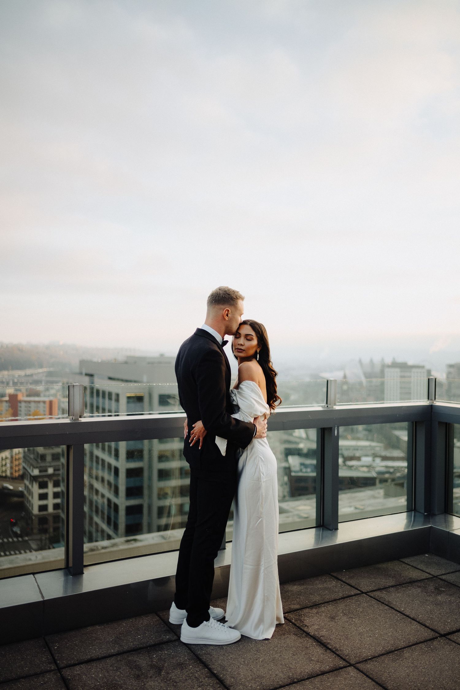 A romantic moment between newlyweds on a high-rise balcony against a misty cityscape.