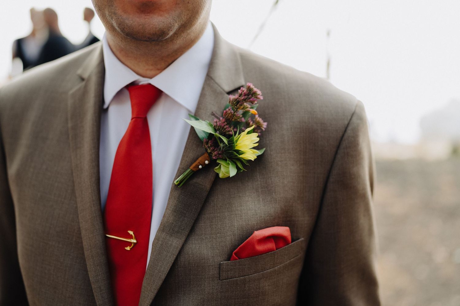 Wedding attire details showing brown suit jacket with bright red tie, gold tie clip and boutonniere with greenery.