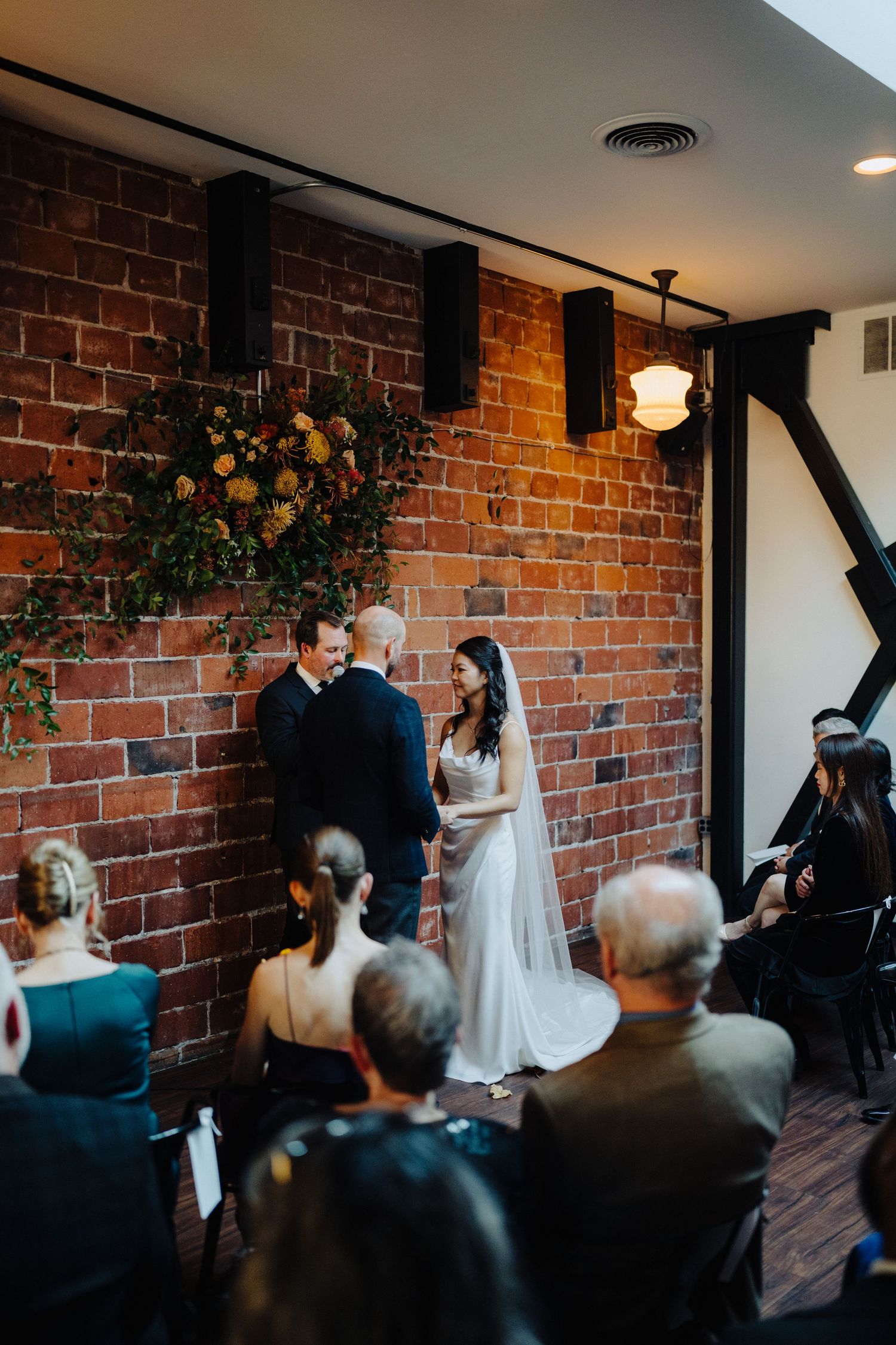 Indoor wedding ceremony in a rustic brick-walled venue with floral decorations.