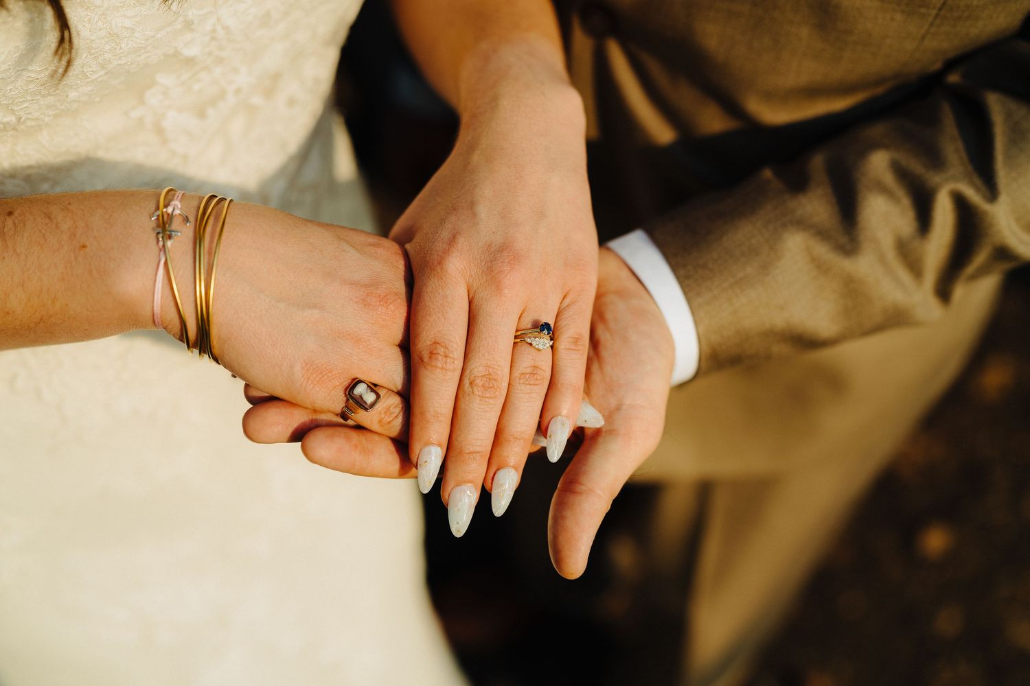 Close-up of hands wearing wedding rings and gold bracelets against white fabric.