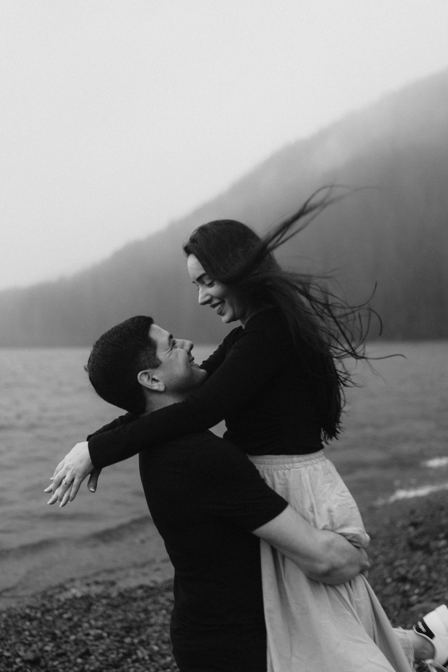 A romantic black and white moment between a couple embracing on a misty beach with mountains in the background.