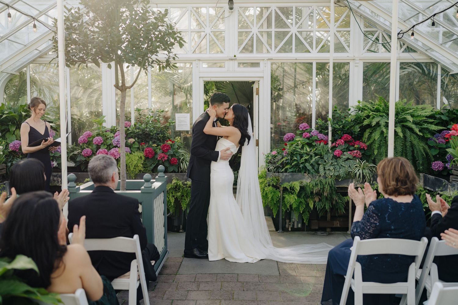 An indoor greenhouse wedding ceremony with guests seated on white chairs among lush plants.
