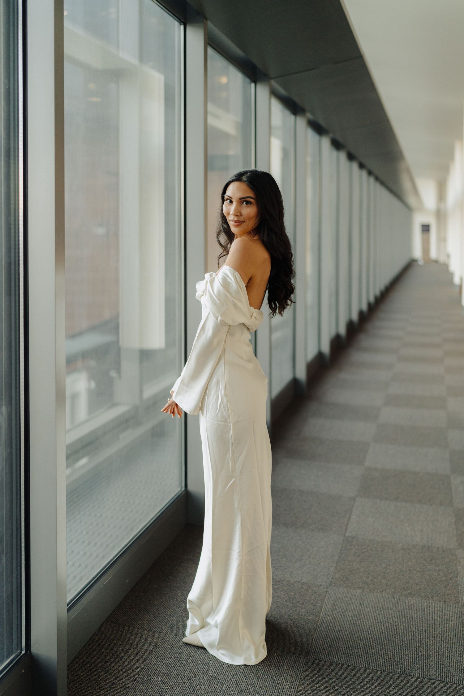 Bride in flowing off-shoulder white gown poses elegantly by window in modern hallway.