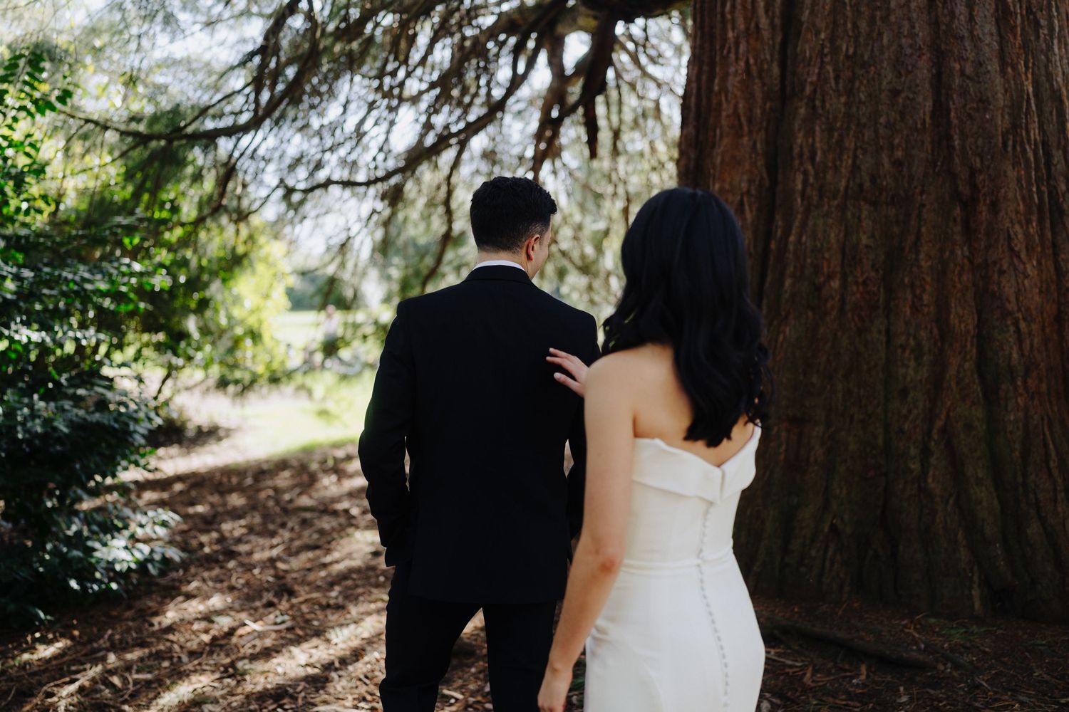 A couple poses under towering redwood trees in formal wedding attire.
