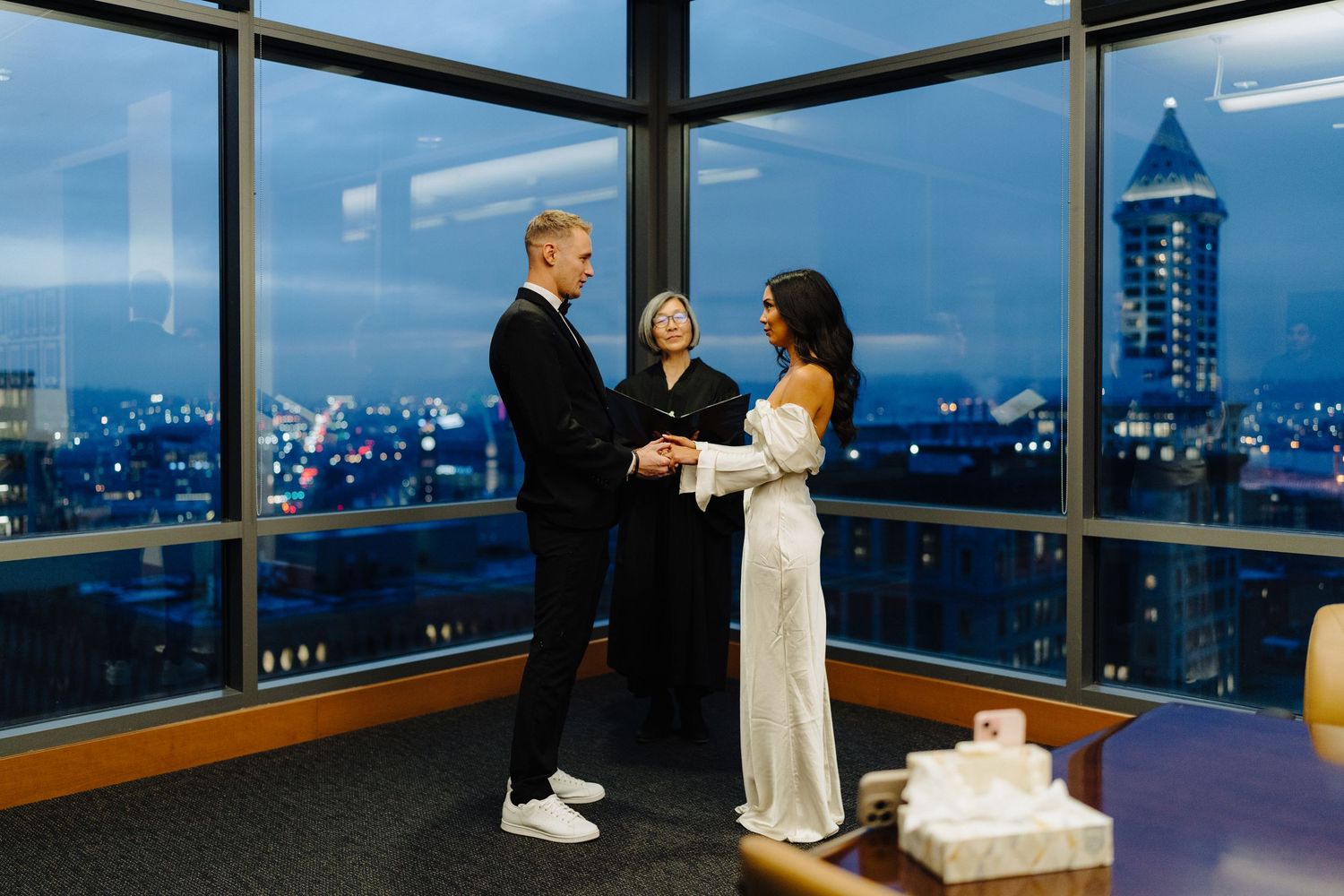 An intimate wedding ceremony takes place in a high-rise venue with city lights visible through windows at dusk.