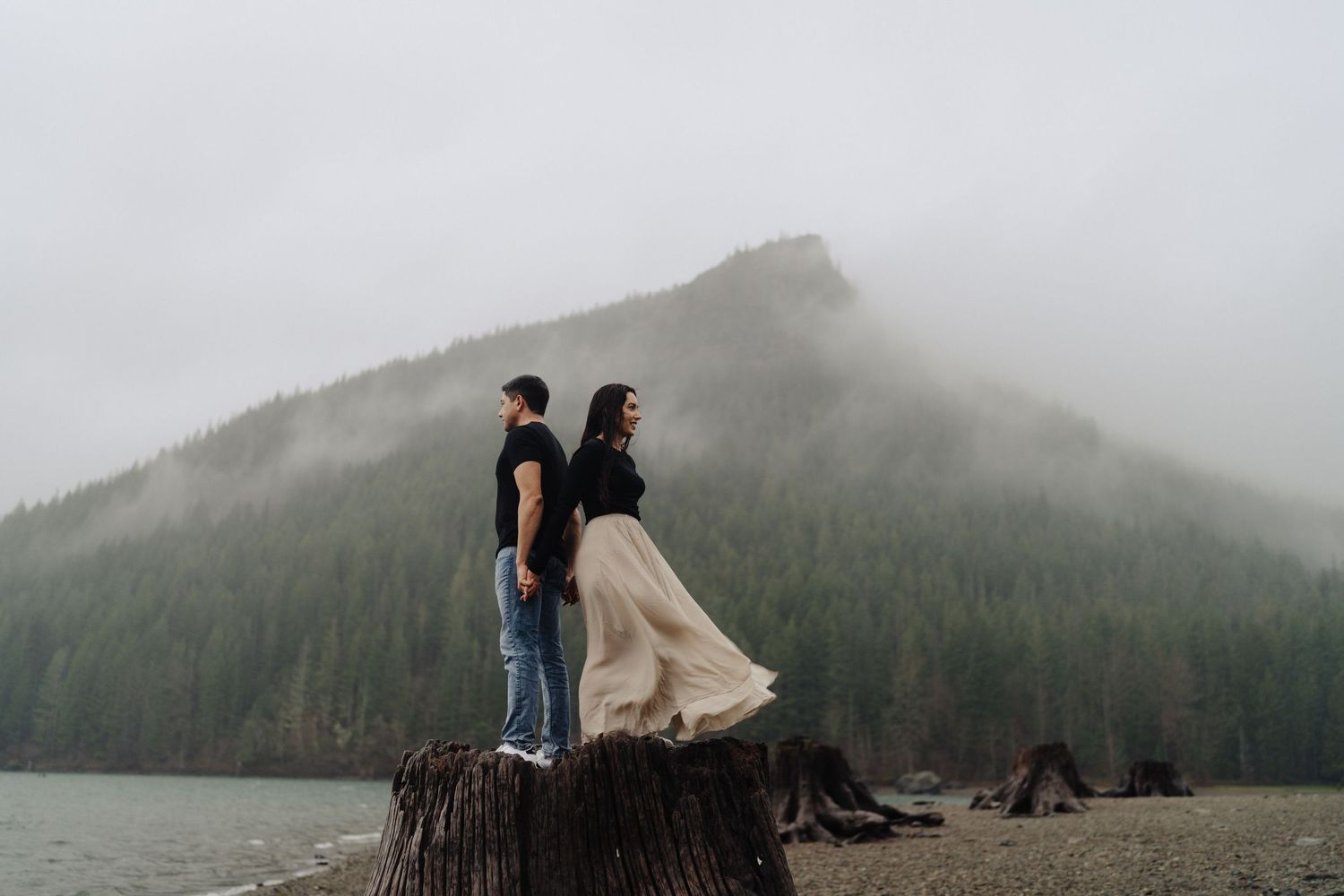 Two people stand on a large tree stump overlooking a misty lake with mountains and forest in the background.