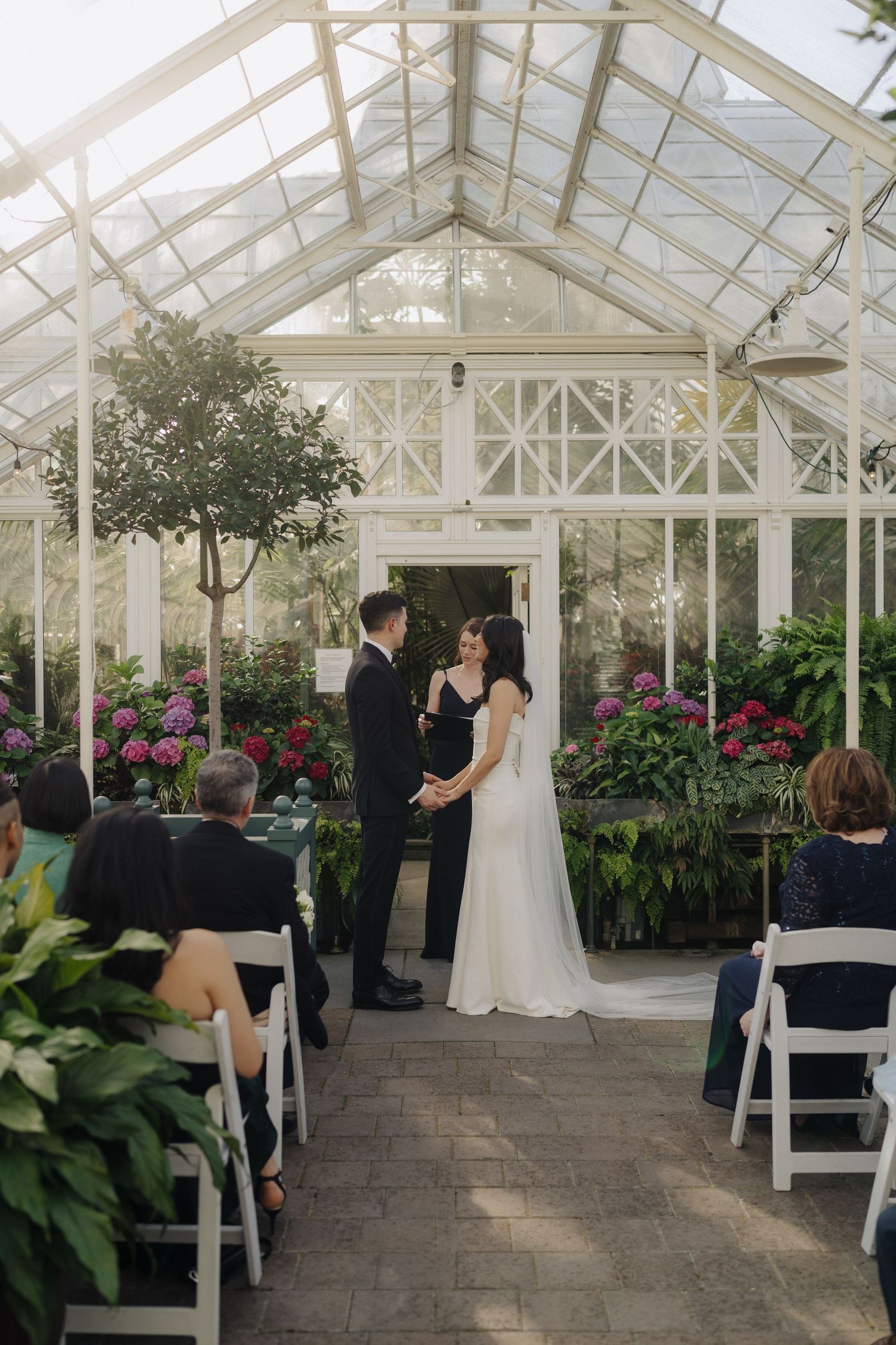 Wedding ceremony taking place in a beautiful greenhouse conservatory filled with colorful flowers.