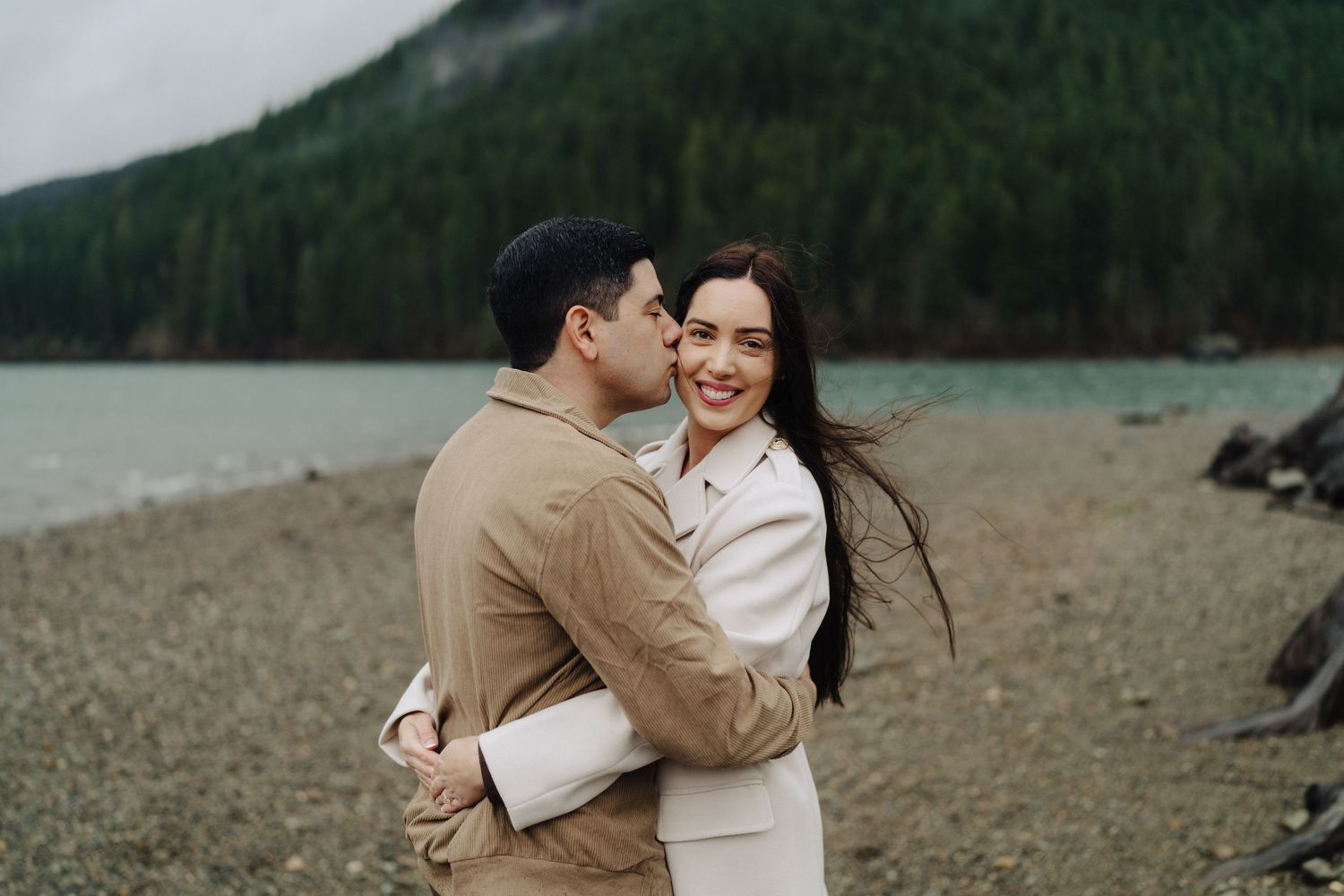 A couple shares a joyful moment on a rocky beach with mountains in the background.