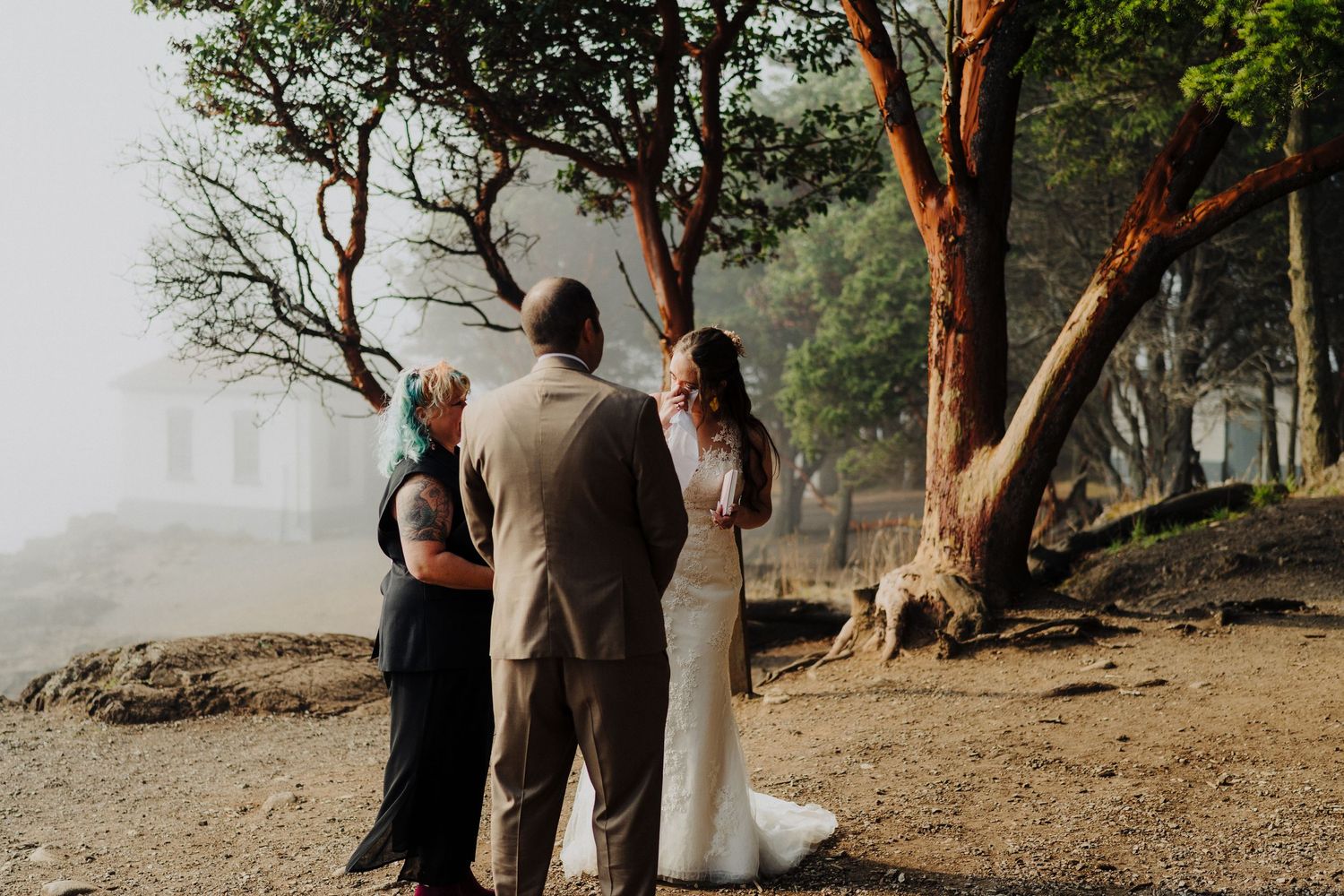 An intimate wedding ceremony takes place under misty trees on a foggy shoreline.