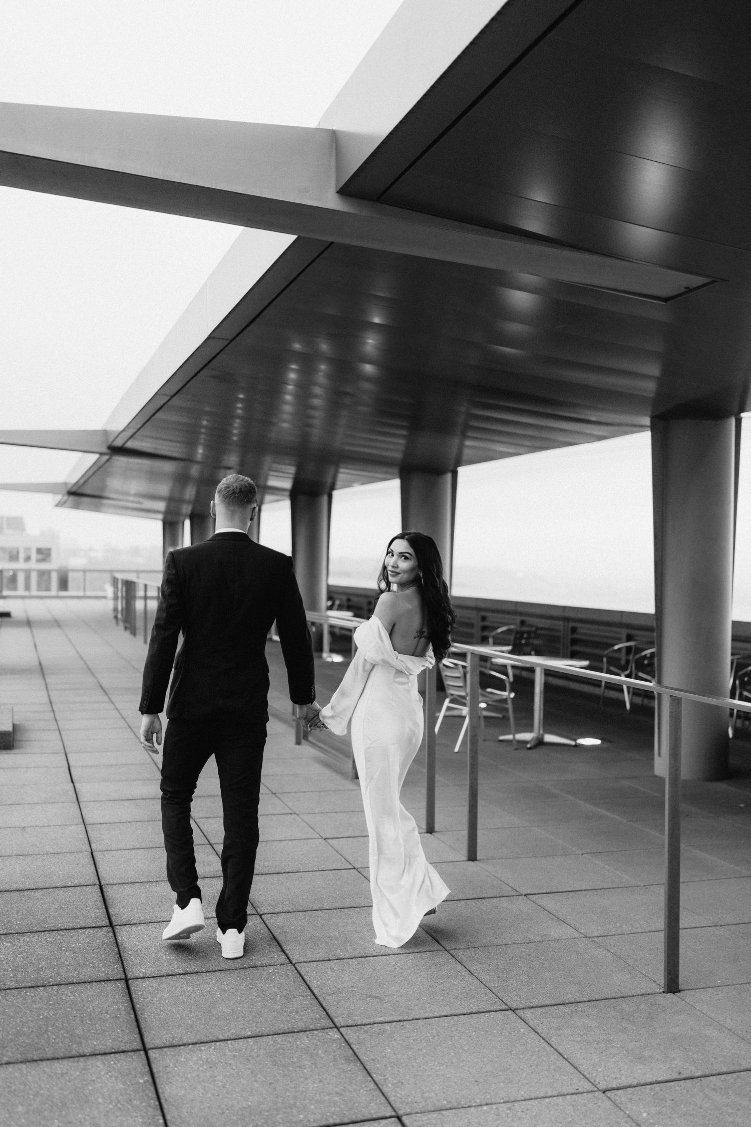 A couple in wedding attire walking together on a modern rooftop terrace in black and white.