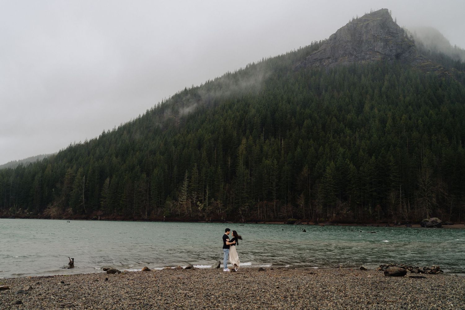 A couple shares an intimate moment on a rocky shoreline with misty mountains and evergreen forest in the background.
