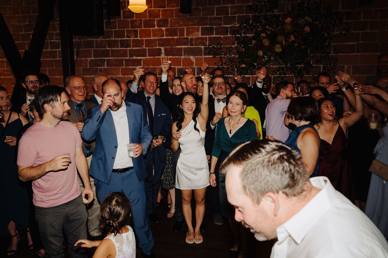 Lively wedding reception crowd dancing and celebrating in front of exposed brick wall.