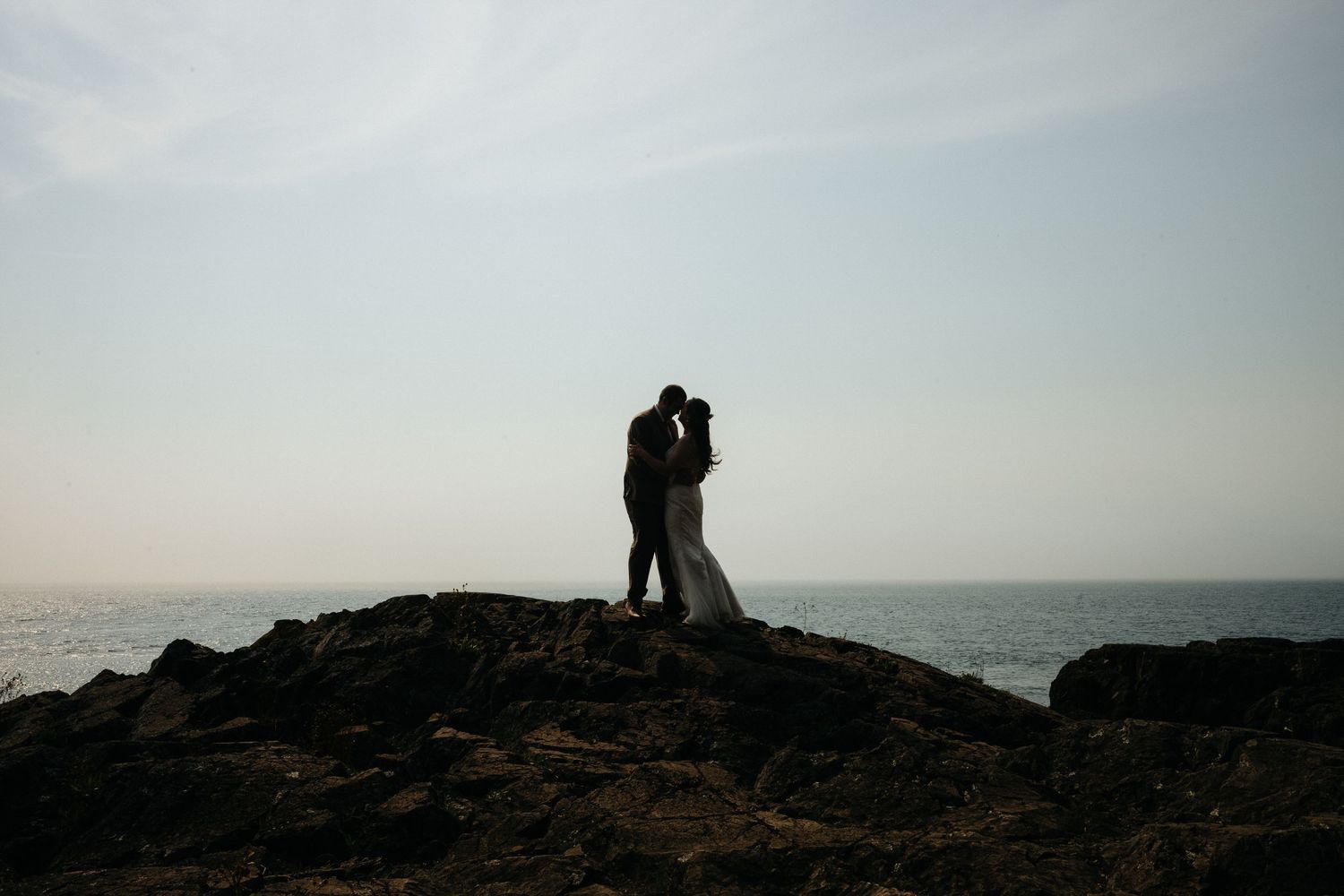 A silhouetted couple stands together on rocky cliffs overlooking the ocean at sunset.