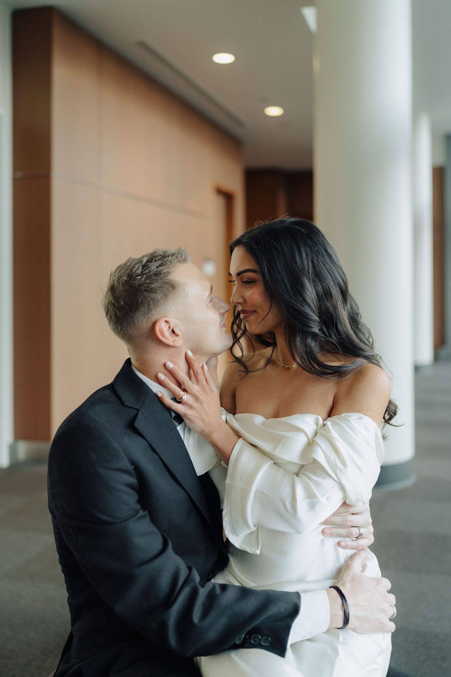 A couple in wedding attire share an intimate moment in an indoor hallway.
