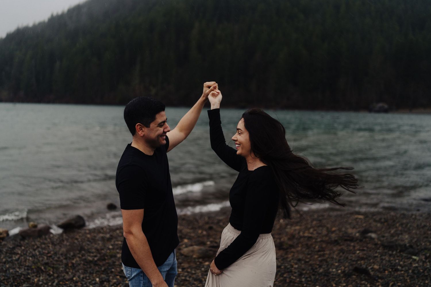 A couple shares an intimate dance moment on a rocky beach with misty mountains and lake in the background.