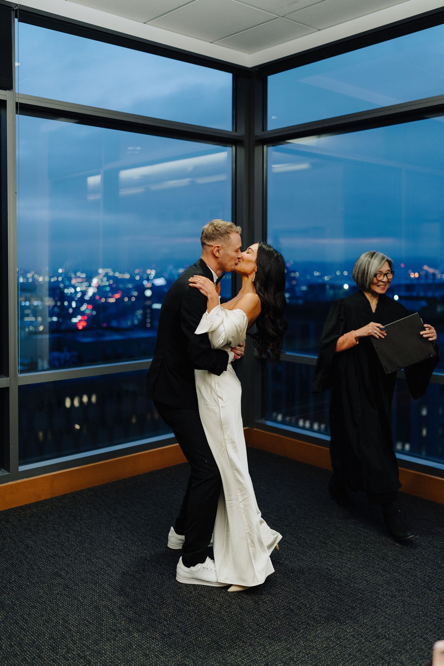 A couple shares their first dance in formal wedding attire against a dramatic cityscape view through floor-to-ceiling windows.