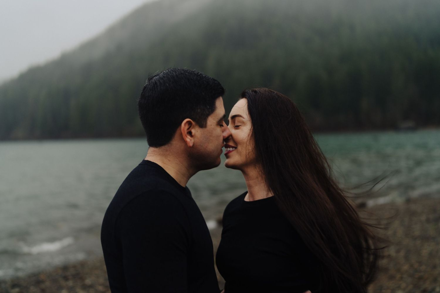 A couple shares an intimate moment by a misty lake with mountains in the background.