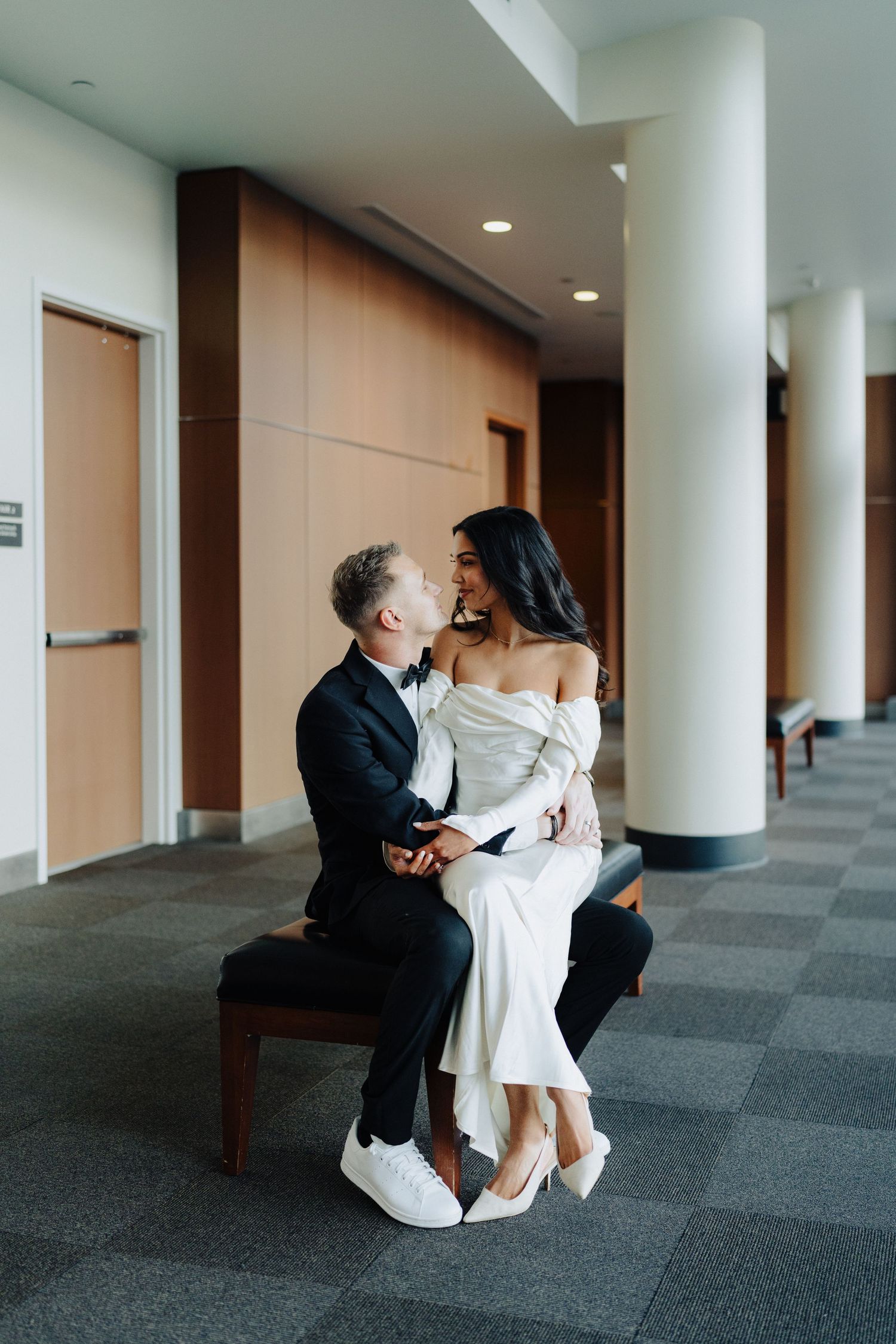 Two people in formal attire share an intimate moment while sitting on a bench in a modern hallway.