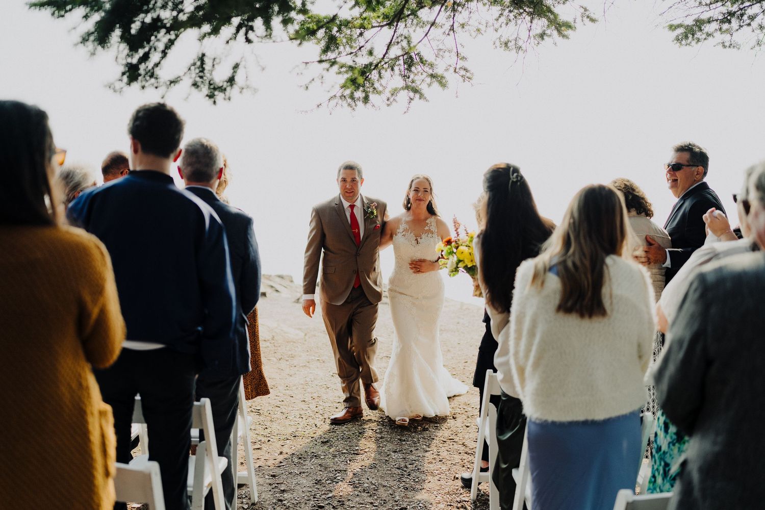 An outdoor wedding ceremony on a sunlit path surrounded by wedding guests in both casual and formal attire.
