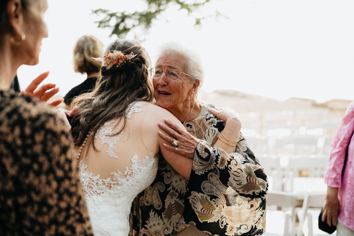 A tender embrace between a bride in lace and an elderly guest at an outdoor wedding celebration.