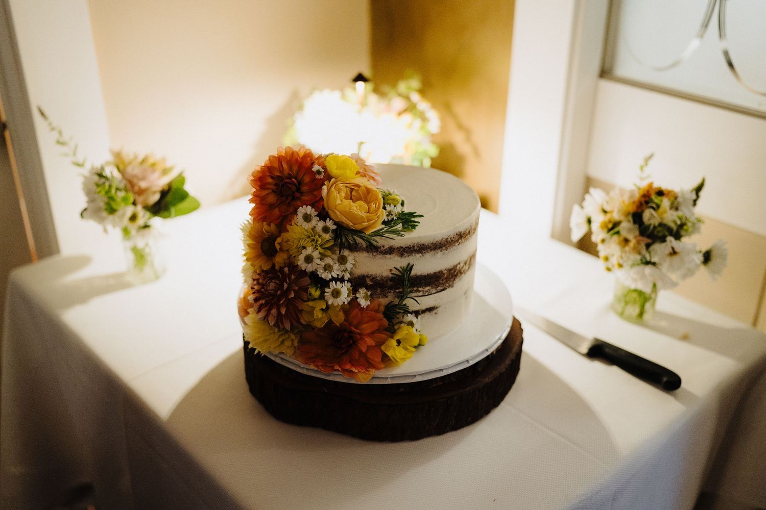 A semi-naked wedding cake decorated with cascading autumn flowers and greenery sits on a white tablecloth.