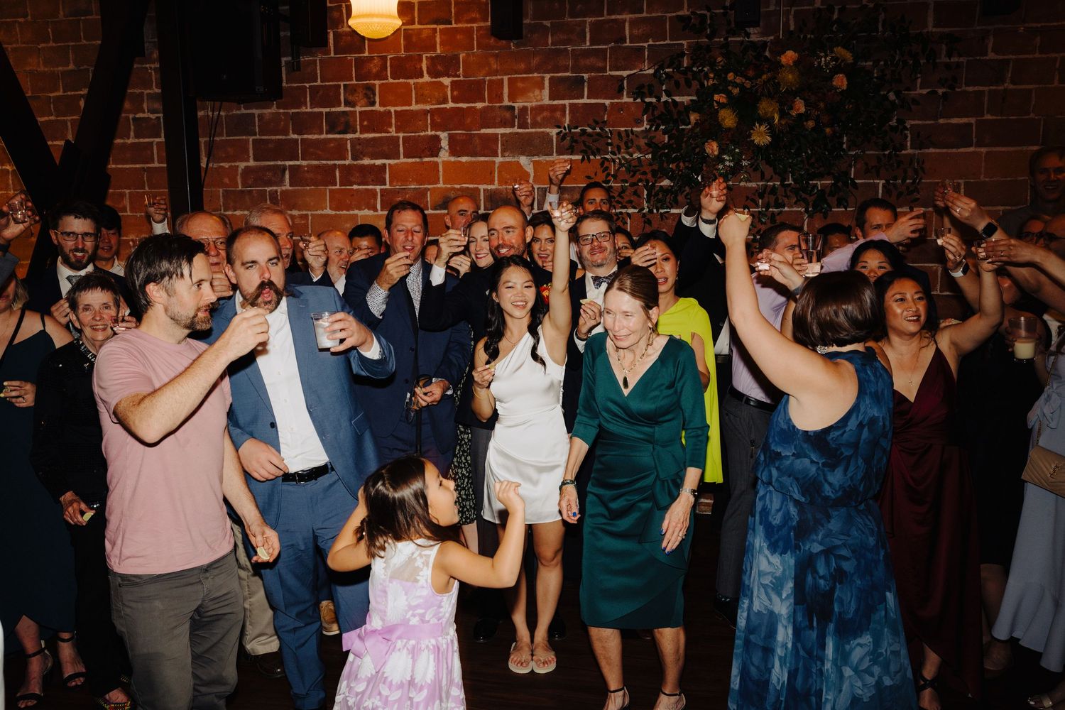 Wedding reception guests dancing and celebrating together in a rustic brick-walled venue.
