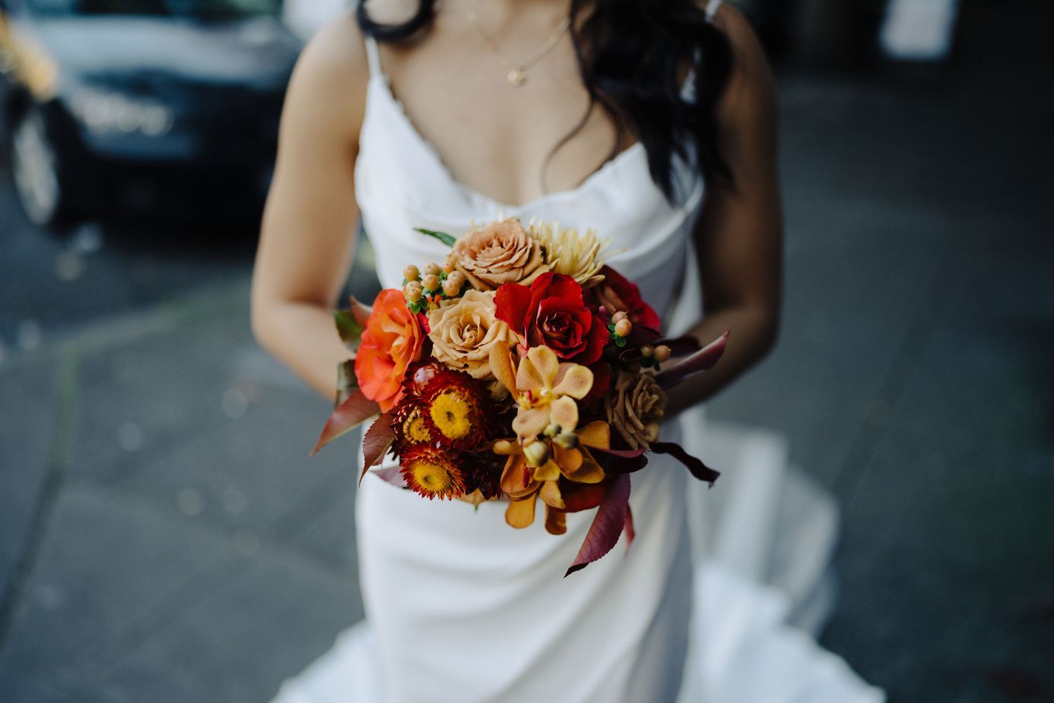 Autumn wedding bouquet with orange and burgundy flowers held against white dress.