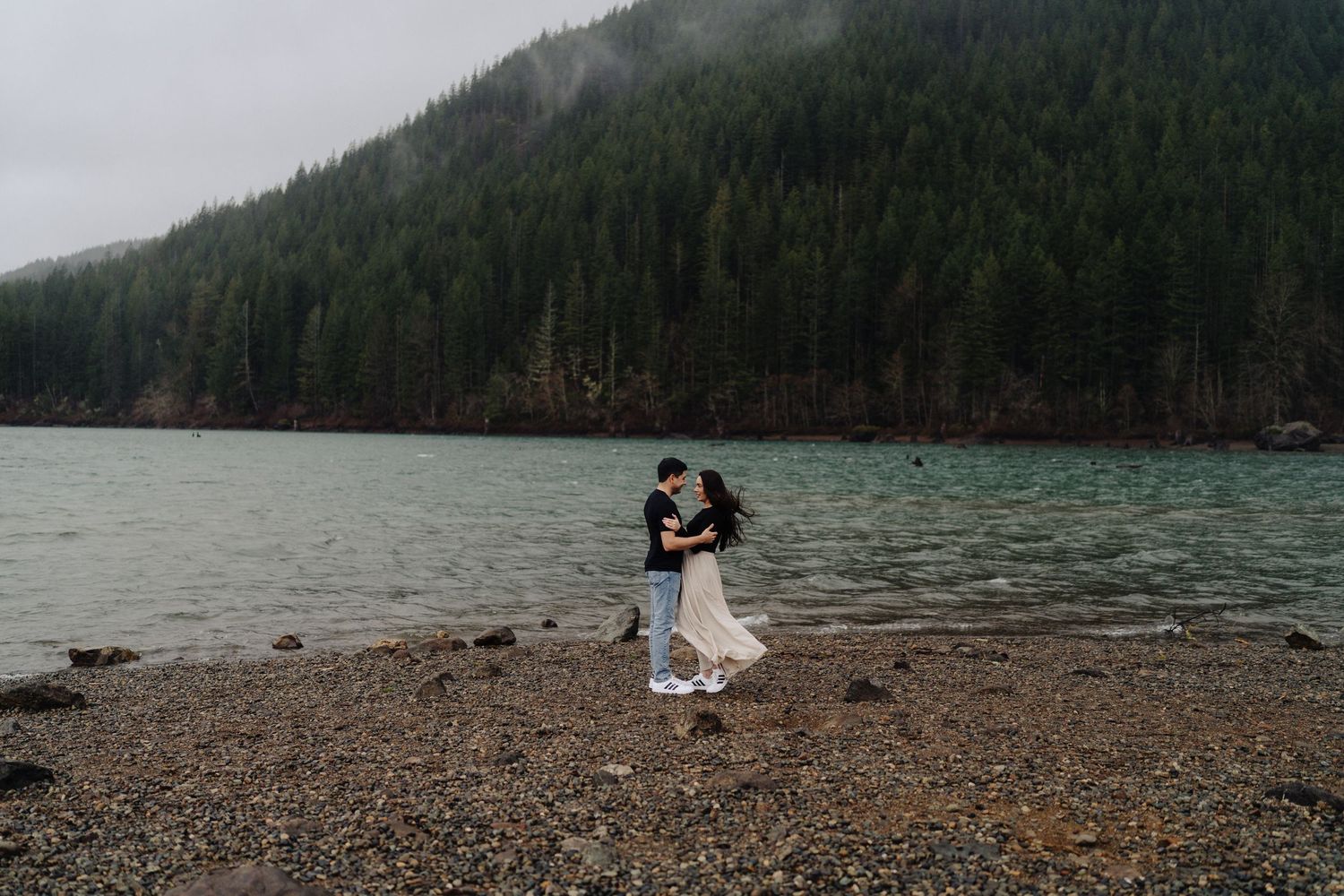 A couple embraces on a pebble beach with misty mountains and evergreen forest behind them.