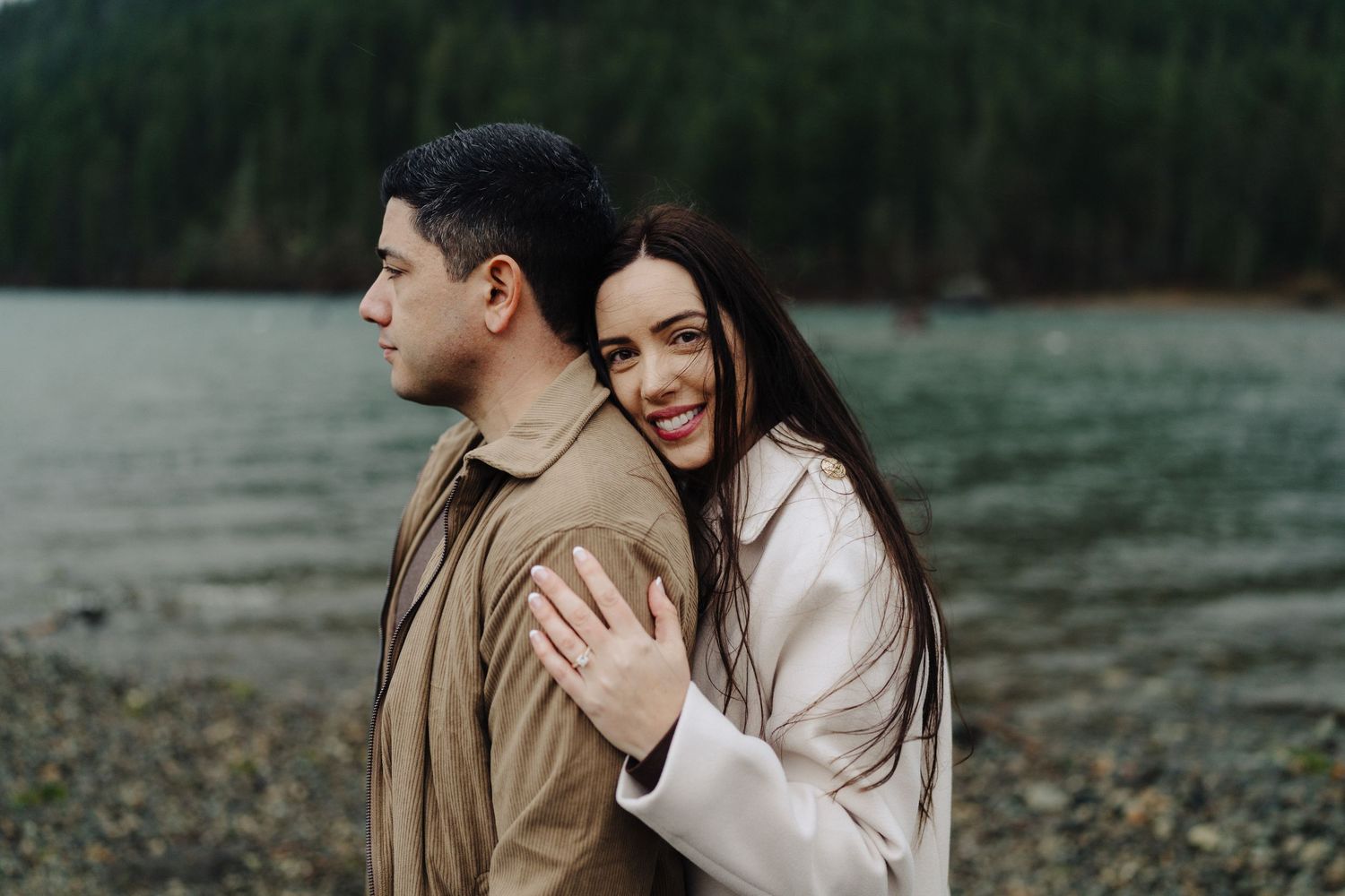 A couple shares an intimate moment on a rocky beach with evergreen mountains and misty lake behind them.