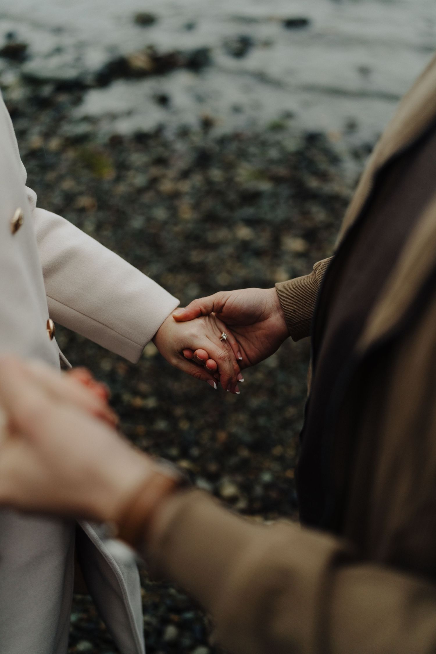 Close-up of hands holding together on rocky beach shore with water in background.
