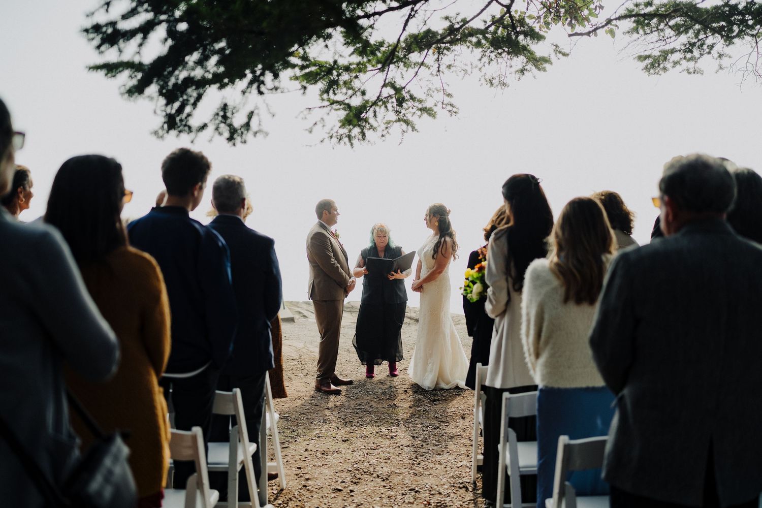 Intimate outdoor wedding ceremony taking place under a tree with guests seated in chairs on a cliff overlooking the ocean.