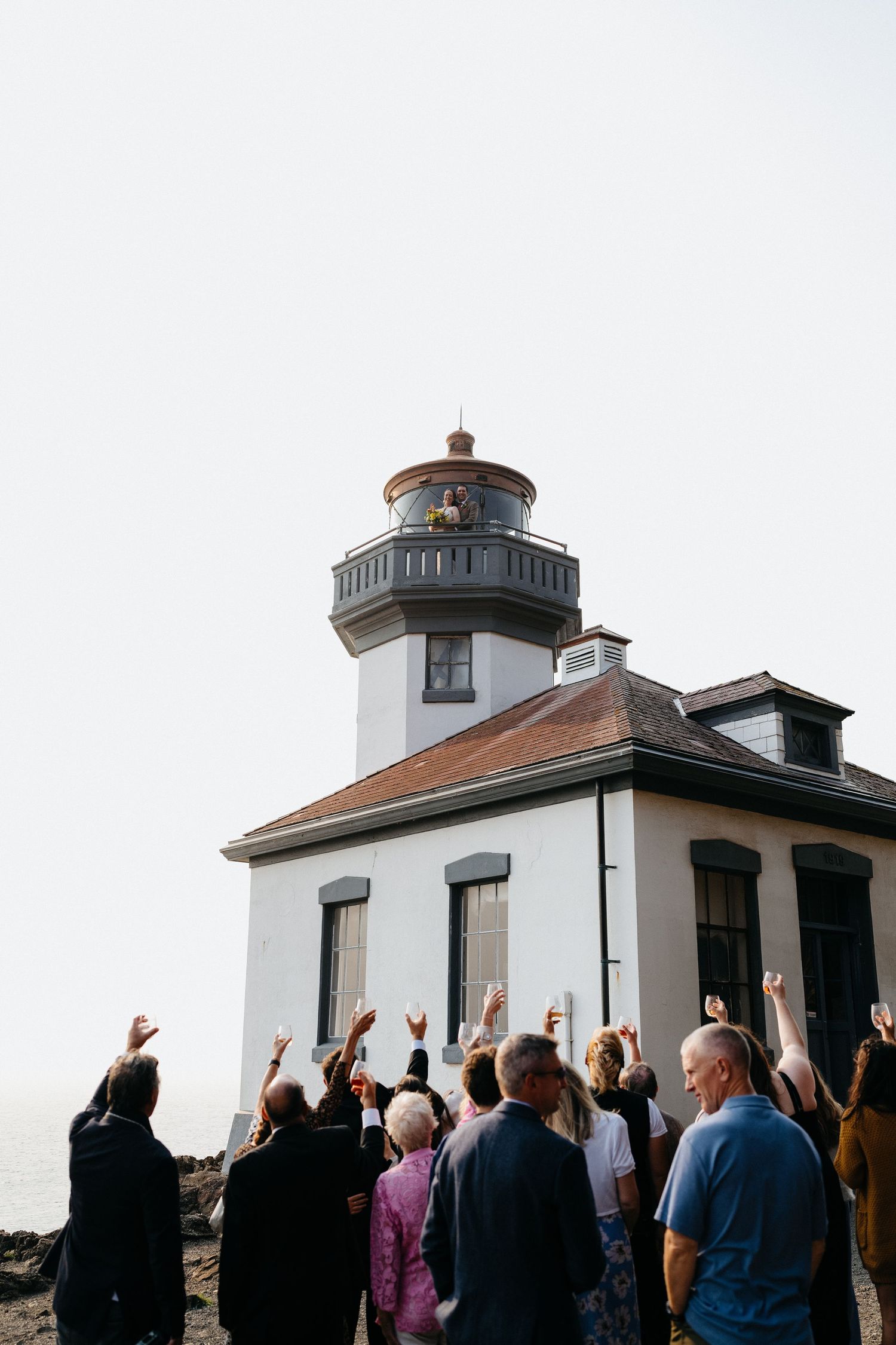 Wedding guests raise their arms in celebration outside a white lighthouse with a brown roof.