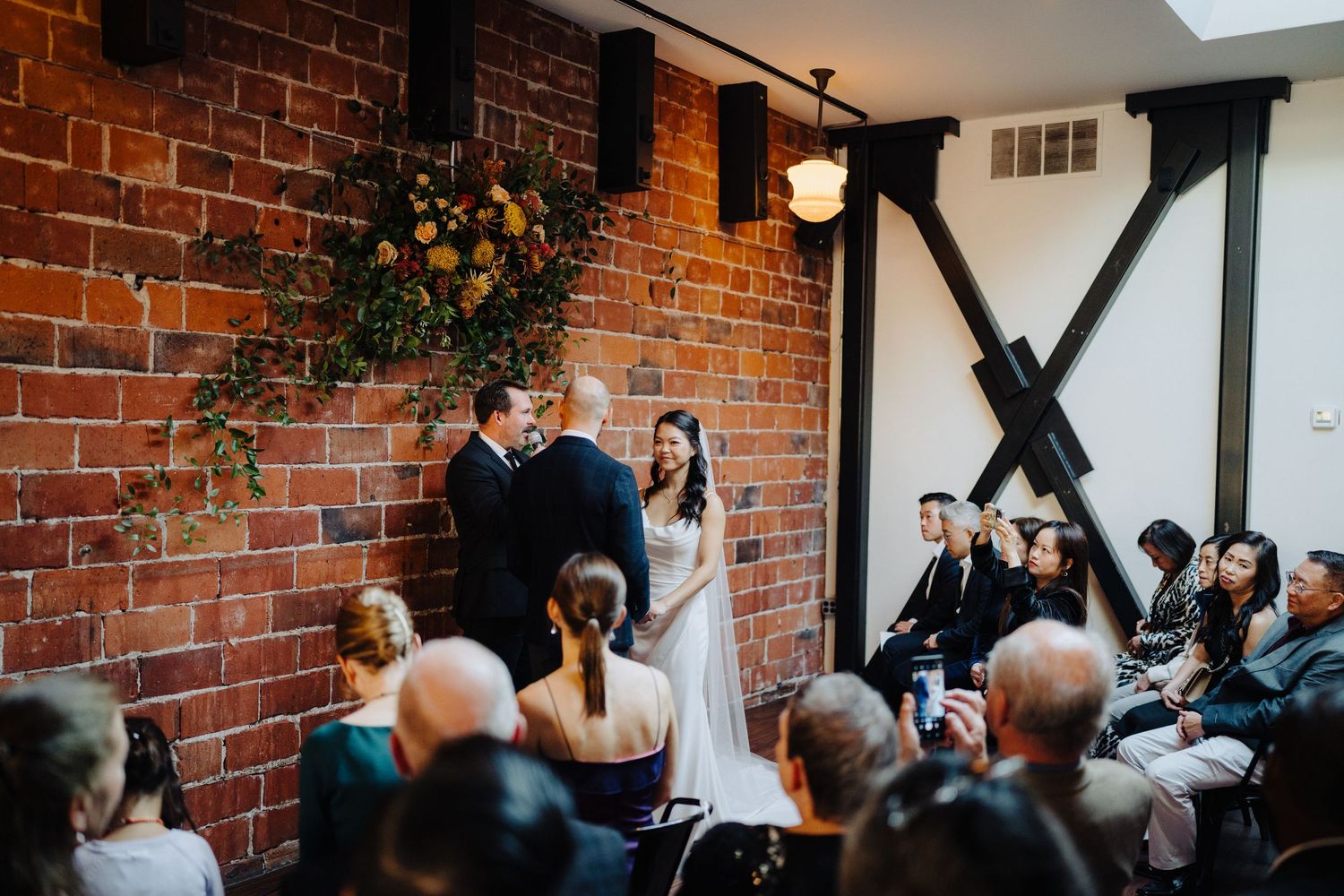 An indoor wedding ceremony in a rustic brick-walled venue with guests seated in rows.